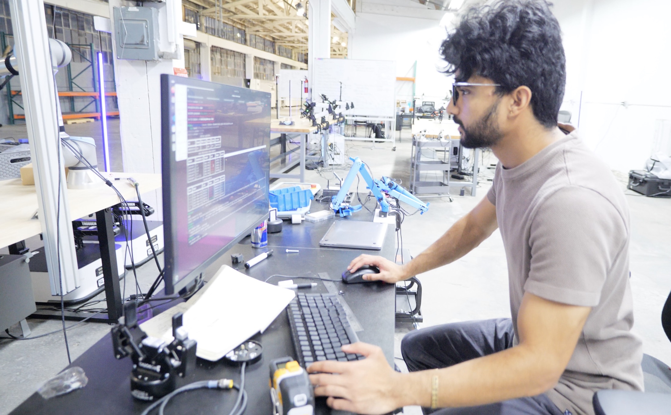 An engineer at a workstation on the Tensr floor, robot arms on the benches behind