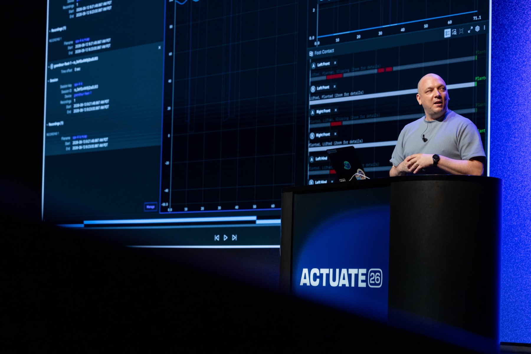 A speaker on the Actuate 26 mainstage in front of a wall-sized robotics data visualization