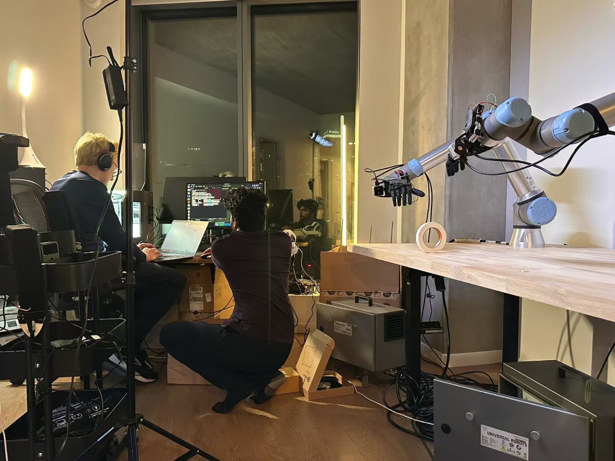 The apartment lab before the factory: a robot arm with a dexterous hand clamped to a desk, two founders working at night
